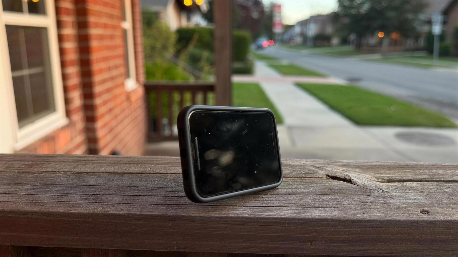 A dark smartphone resting on a wooden porch railing with a suburban street softly blurred behind it